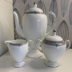 Könnte beinhalten: Ein weißes Porzellan-Teeservice mit silbernen Verzierungen, bestehend aus einer Kaffeekanne, einem Sahnekännchen und einer Zuckerdose. Die Kaffeekanne hat ein hohes, schlankes Design mit gebogenem Ausguss und silbernem Deckel. Sahnekännchen und Zuckerdose haben passende silberne Akzente.