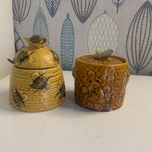 Op de afbeelding: Twee keramische honingpotten. Eén is in de vorm van een bijenkorf met een bij op het deksel en een lepel. De andere is een ronde pot met een zonnebloemmotief en een bij op het deksel. Beide zijn bruin en geel.