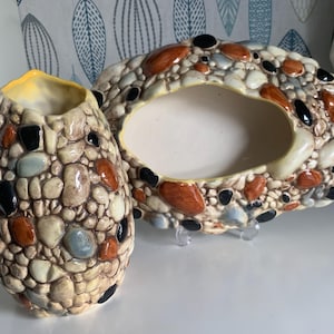 Peut inclure: Deux pots en céramique en forme de galets, l'un est un vase et l'autre un bol. Les deux sont décorés d'un motif de galets dans des tons de brun, beige, gris et noir.