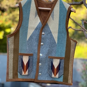 May include: A blue, brown, and white patterned vest with a suede trim and two pockets. The vest has a button closure and is hanging on a wooden hanger.