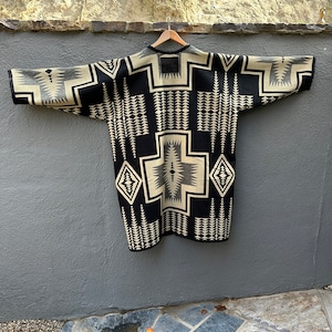May include: A black and cream patterned Pendleton wool blanket with a geometric design. The blanket is hanging on a hanger against a gray wall.