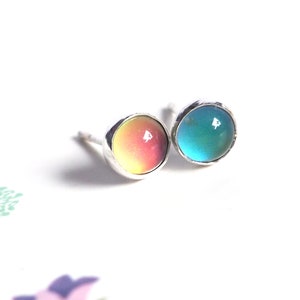 Puede incluir: Un par de pendientes de botón de plata con piedras preciosas redondas e iridiscentes. Una piedra preciosa es un degradado de amarillo a rosa, y la otra es de color azul verdoso.