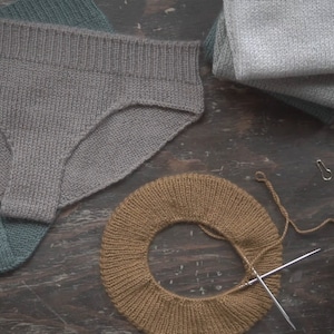 Puede incluir: Primer plano de tres artículos de punto: una braga gris, un gorro de punto marrón y una bufanda gris. Los artículos están dispuestos sobre una superficie de madera.