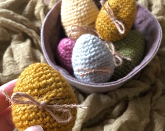Può includere: Una collezione di uova di Pasqua all'uncinetto in un cestino. Le uova sono di vari colori, tra cui giallo, verde, rosa e grigio. Ogni uovo è legato con un fiocco di spago marrone.