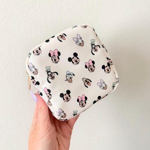 Puede incluir: Un pequeño neceser cuadrado con fondo color crema. La bolsa está cubierta con un patrón repetido de caras de personajes de dibujos animados, incluyendo Mickey Mouse, Minnie Mouse, Donald Duck y Daisy Duck. La bolsa tiene una cremallera dorada.