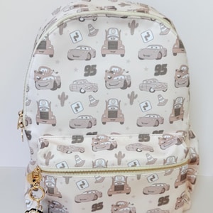 May include: A cream-coloured backpack featuring an all-over print of cartoon cars, traffic cones, cacti, and the number 95. The bag includes a front zipped pocket with a gold-toned zip and a dangling charm shaped like a car character.