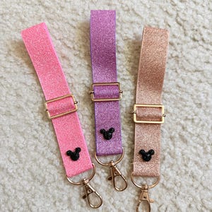 May include: Three glittery keychains in pink, purple, and rose gold. Each keychain features a gold-colored buckle and clasp, with a small black mouse silhouette charm. The keychains are displayed on a white, textured surface.