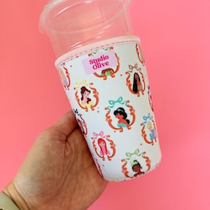 Könnte beinhalten: Ein durchsichtiger Kunststoffbecher mit einer weißen Stoffhülle, die ein wiederkehrendes Muster verschiedener Zeichentrickprinzessinnen in verzierten goldenen Rahmen zeigt. Ein rosa Etikett mit dem Text Studio Olive ist oben angebracht.