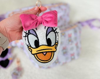 May include: A plush Daisy Duck keychain with a pink bow. The keychain features Daisy's face with embroidered details, including black eyelashes, purple eyelids, and a yellow beak. A gold-colored ring is attached to the top.