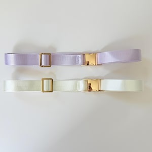 May include: Two adjustable belts. One is a pale lavender color, and the other is white. Both belts have gold-colored metal buckles and rectangular belt loops. The belts are made of a smooth, woven material.