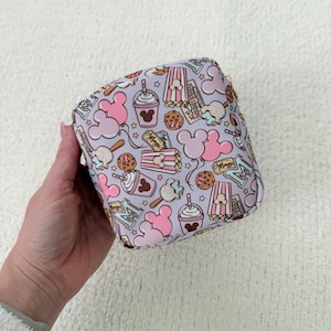May include: A small, square cosmetic bag with a repeating pattern of pink Mickey Mouse heads, popcorn, cookies, and drinks on a light purple background. The bag has a gold zipper and is held in a hand.