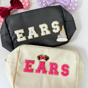 Puede incluir: Dos bolsas blancas con cremallera con la palabra "EARS" en letras doradas y rosas. Las bolsas tienen diferentes adornos, una con un castillo y la otra con una cabeza de Minnie Mouse.