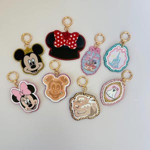 Könnte beinhalten: Eine Sammlung von acht Schlüsselanhängern mit verschiedenen Charakter-Motiven, darunter Mickey Mouse, Minnie Mouse, eine Waffel, ein Auto, ein Schloss und ein Geist, jeweils befestigt an einem goldfarbenen Metallring zum Aufhängen.