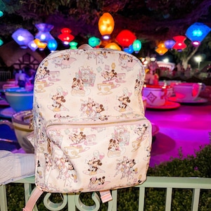 May include: A white backpack with a repeating pattern of Minnie Mouse and Mickey Mouse in teacups and other whimsical designs. The backpack has a zippered main compartment, a front pocket, and pink straps. The background features colorful lights.
