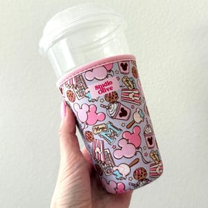 May include: A clear plastic tumbler with a pink and lavender patterned sleeve featuring Mickey Mouse icons, popcorn, and ice cream. The sleeve has the text "Studio Olive". The tumbler has a clear lid.