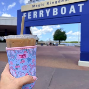 May include: A clear plastic cup with iced coffee and a straw, held in a light blue cup sleeve with a pink top. The sleeve has a pattern of castles, bows, and roses. The text "Studio Olive" is visible on the sleeve. The background shows a blue sign that says "Magic Kingdom FERRYBOAT".