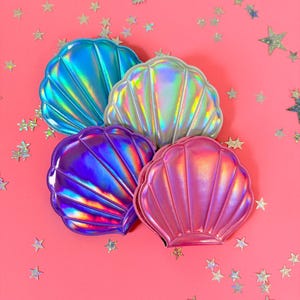 Puede incluir: Cuatro estuches cosméticos con forma de concha iridiscente en azul, plateado, morado y rosa sobre un fondo rosa con estrellas de purpurina plateadas.
