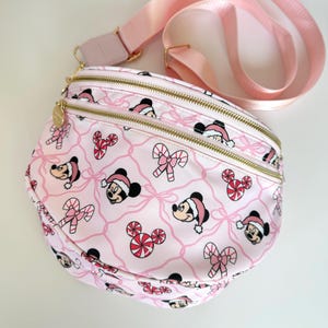 May include: A light pink crossbody bag featuring a repeating pattern of Mickey Mouse heads wearing Santa hats, candy canes, and peppermint swirls. The bag has two gold-tone zippered compartments and an adjustable light pink nylon shoulder strap.