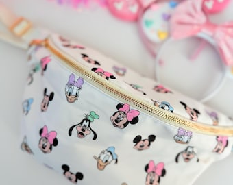 May include: A white fanny pack with a gold zipper and a pattern of various Disney characters, including Mickey Mouse, Minnie Mouse, Donald Duck, Goofy, and Pluto. The characters are in various colors, including pink, blue, yellow, and green.