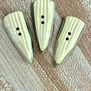 May include: Three decorative, shield-shaped buttons with a light yellow, striped design. Each button has two black holes for sewing. The buttons have a light green and gray border and are arranged on a light brown, wood-grain surface.