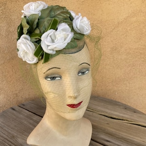 Puede incluir: Un sombrero blanco con velo verde y rosas de tela blanca. El sombrero está en una cabeza de maniquí.