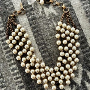 Puede incluir: Un collar de perlas vintage con múltiples hilos de perlas de imitación. El collar presenta una cadena y un cierre dorados. Las perlas son de color blanco roto y están dispuestas en un diseño en cascada, creando un efecto de capas. El collar se muestra sobre una superficie con estampado gris y blanco.