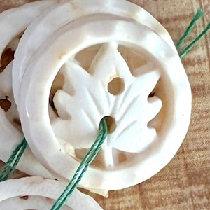 May include: Close-up of several off-white, circular beads with a carved leaf design in the center. Each bead has a hole for stringing, and green string is visible. The beads are arranged on a light brown surface.