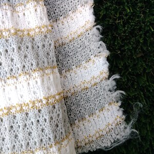 May include: White and gray knitted fabric with gold thread woven throughout. The fabric has a fringed edge.