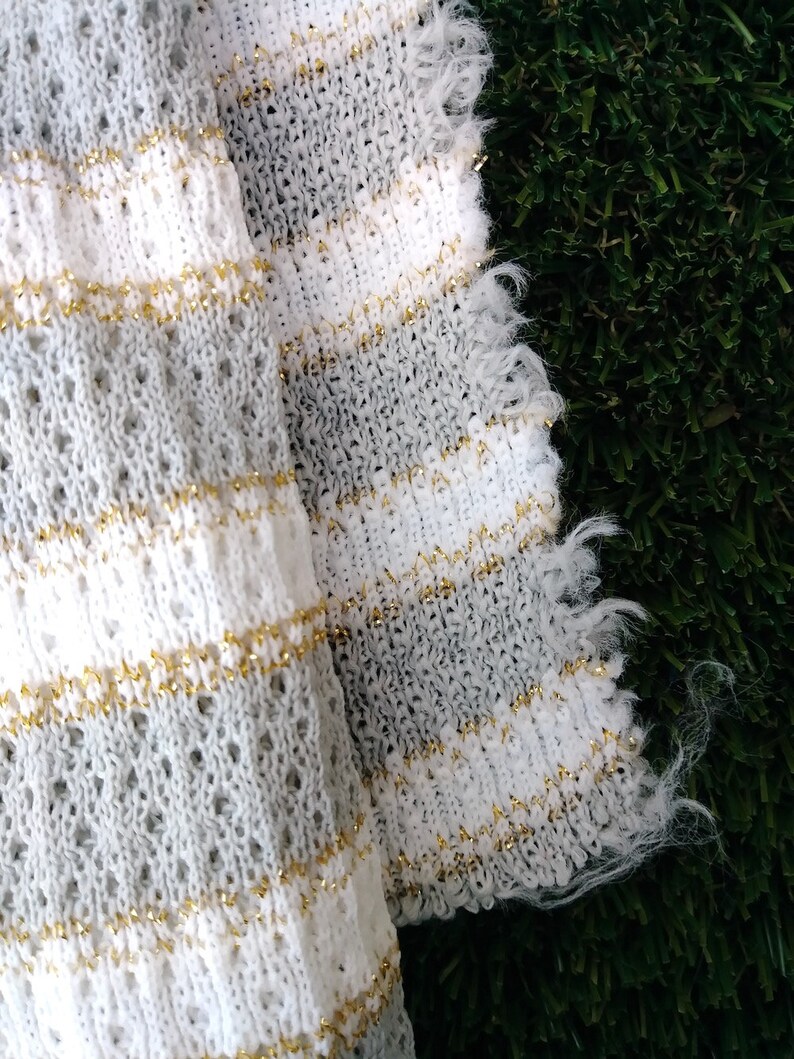 May include: White and gray knitted fabric with gold thread woven throughout. The fabric has a fringed edge.