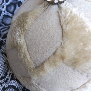Puede incluir: Un sombrero de piel sintética beige con un diseño acolchado y un adorno de diamantes de imitación en la parte superior.