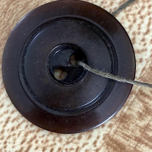 May include: A dark brown, circular button with two holes and a textured surface. A length of gray thread is threaded through the button. The button has a raised, concentric ring design.