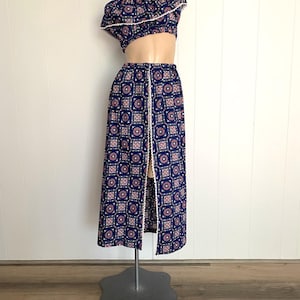 May include: A two-piece vintage outfit featuring a one-shoulder crop top and a long skirt. The set is navy blue with a repeating geometric pattern in pink and white. The edges are trimmed with white lace.