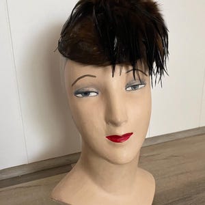 Puede incluir: Un sombrero vintage de plumas marrones expuesto en una cabeza de maniquí. El sombrero tiene una base marrón oscuro con un grupo de plumas en la parte superior. El maniquí tiene rasgos pintados, incluyendo labios rojos y sombra de ojos azul.