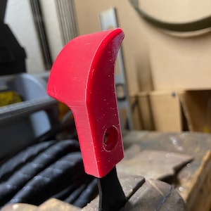 Może przedstawiać: Czerwony plastikowy uchwyt z czarnym metalowym ostrzem, zaciśnięty w imadle.