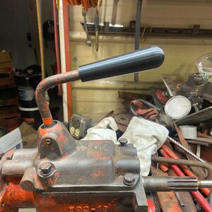 May include: Close-up of a rusty orange metal gear with a black plastic handle. The handle is bent into a loop shape and is attached to the gear. The gear is surrounded by various tools and other metal objects.