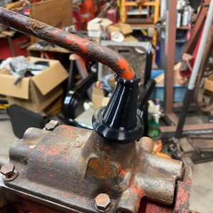 May include: Close-up of a rusty orange metal gear shifter with a black rubber boot. The shifter is attached to a larger metal gear box.