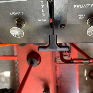 Könnte beinhalten: Nahaufnahme eines schwarzen Steuerhebels mit T-förmigem Griff. Der Hebel befindet sich auf einer grauen Steuertafel mit dem Wort "LIGHTS". Weitere Bedienelemente sind "FRONT P.T.O." und "SLOW". Der Hintergrund ist eine rot-orange Maschine.