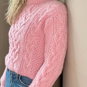 Puede incluir: Un suéter de punto de cable rosa con cuello alto y mangas largas.