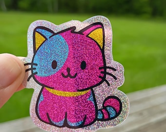 Op de afbeelding: Een holografische sticker van een cartoonkat met een roze lichaam, blauwe en roze kop en gele oren. De kat heeft een lachend gezicht en een gele kraag. De sticker heeft een glinsterende, iriserende afwerking.