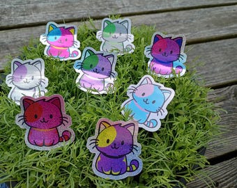 Op de afbeelding: Een verzameling kleurrijke kattenstickers met holografische accenten. Elke sticker heeft een ander kleurenschema, waaronder roze, blauw, paars en groen. De stickers zijn gerangschikt op een bed van groen gebladerte tegen een houten achtergrond.