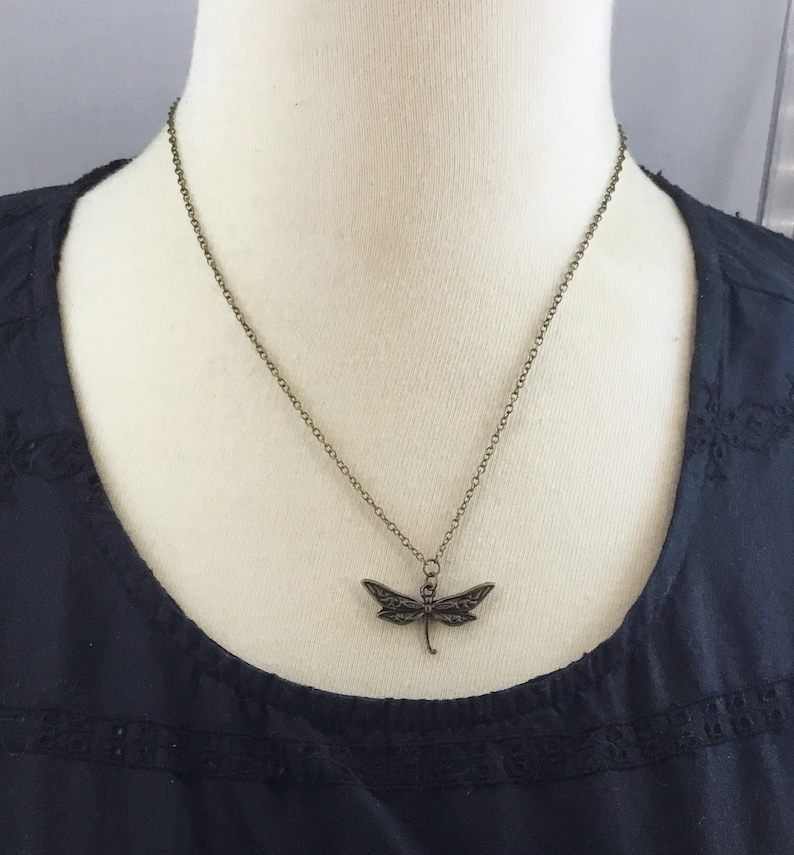 Peut inclure: Un collier en cha&icirc;ne bronze avec un pendentif en forme de libellule. La libellule est d&eacute;taill&eacute;e avec des ailes et un corps complexes.