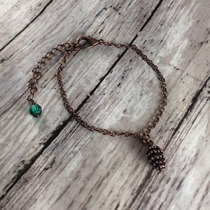 May include: A copper chain bracelet with a small pine cone charm and a green glass bead. The bracelet has a lobster clasp closure.