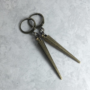 May include: A pair of bronze-colored earrings with a pointed design. The earrings are made of metal and have a simple, minimalist style.