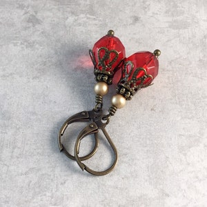 May include: A pair of red glass earrings with antique gold filigree accents and pearl beads. The earrings have a teardrop shape and are suspended from leverback closures.