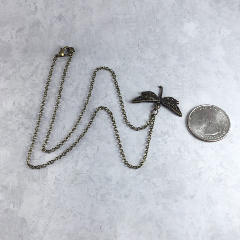 Peut inclure: Un collier en cha&icirc;ne bronze avec un pendentif en forme de libellule. Le pendentif est d&eacute;taill&eacute; et complexe, avec des ailes d&eacute;licates et un corps long et fin. La cha&icirc;ne est une cha&icirc;ne simple et li&eacute;e.