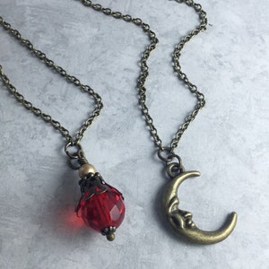 May include: Two necklaces with bronze chains and pendants. One pendant is a red faceted glass teardrop with a bronze filigree top. The other pendant is a bronze crescent moon with a face.