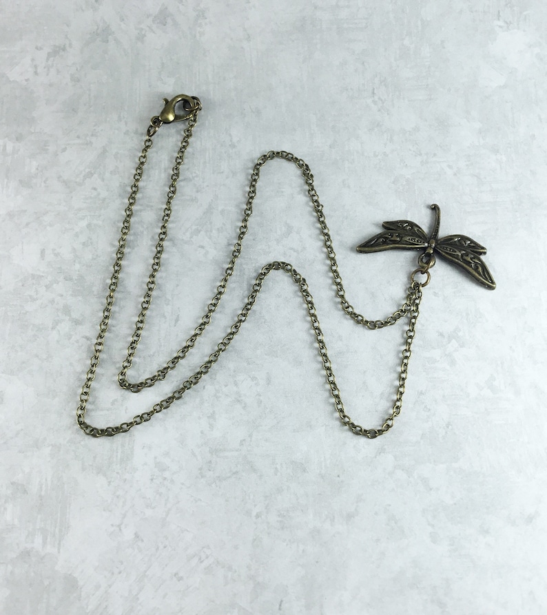 Peut inclure: Un collier en cha&icirc;ne bronze avec un pendentif d&eacute;licat en forme de libellule. La libellule pr&eacute;sente des d&eacute;tails complexes et une finition l&eacute;g&egrave;rement oxyd&eacute;e.