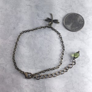 May include: A delicate bronze chain bracelet featuring a dragonfly charm and a green glass bead. The bracelet has a lobster clasp fastening.
