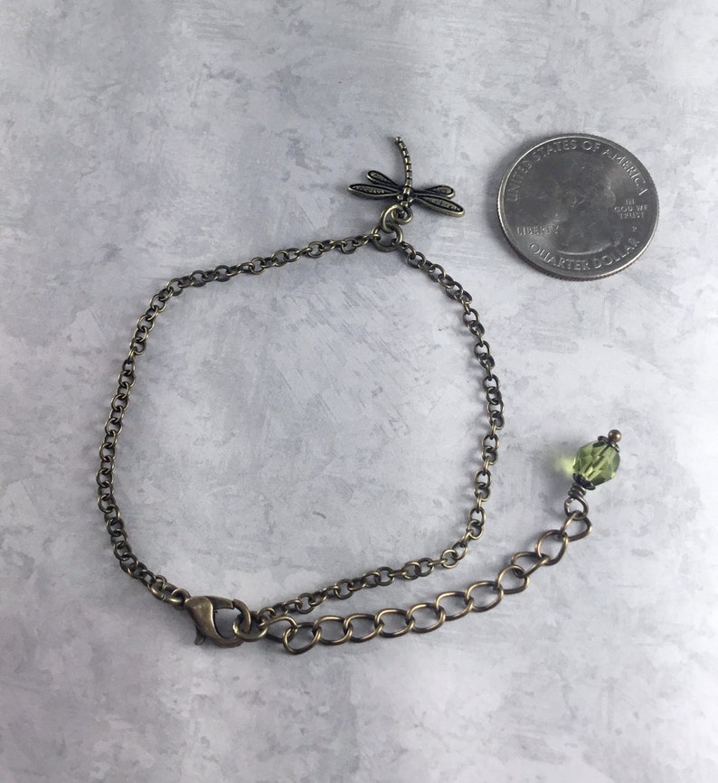 May include: A delicate bronze chain bracelet featuring a dragonfly charm and a green glass bead. The bracelet has a lobster clasp fastening.