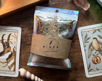 Könnte beinhalten: Nahaufnahme eines kleinen, durchsichtigen Plastikbeutels, gefüllt mit einer Mischung aus getrockneten Kräutern und rosa Himalaya-Salz. Der Beutel ist mit "Pine & Pestle, House of Herbals" beschriftet. Im Hintergrund sind zwei Tarotkarten zu sehen, eine zeigt ein Antilopen und Weizen, die andere einen Finken und einen Pfirsich.